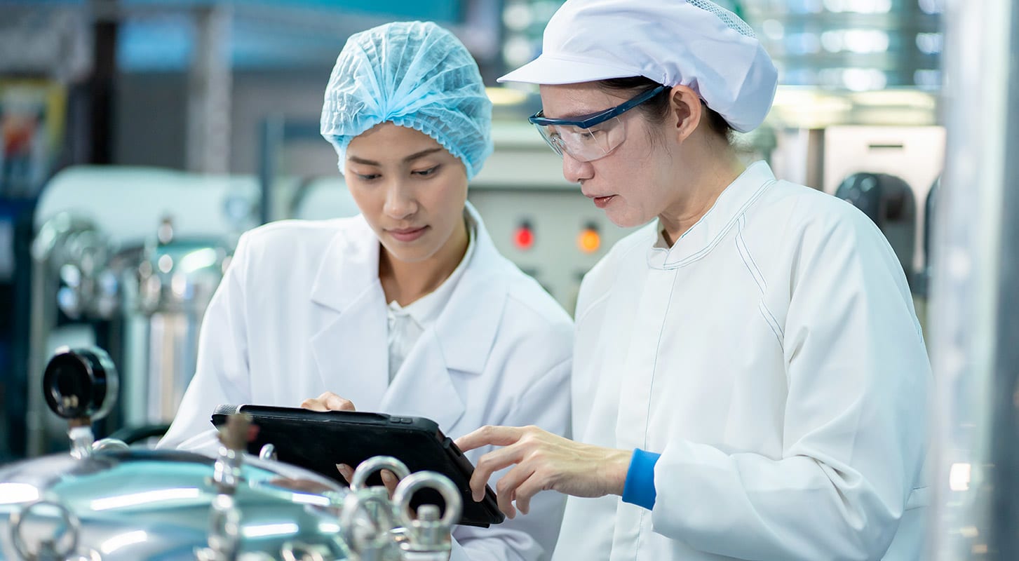 Two female scientists in white lab coats reviewing data on a tablet, collaborating in a food development facility.